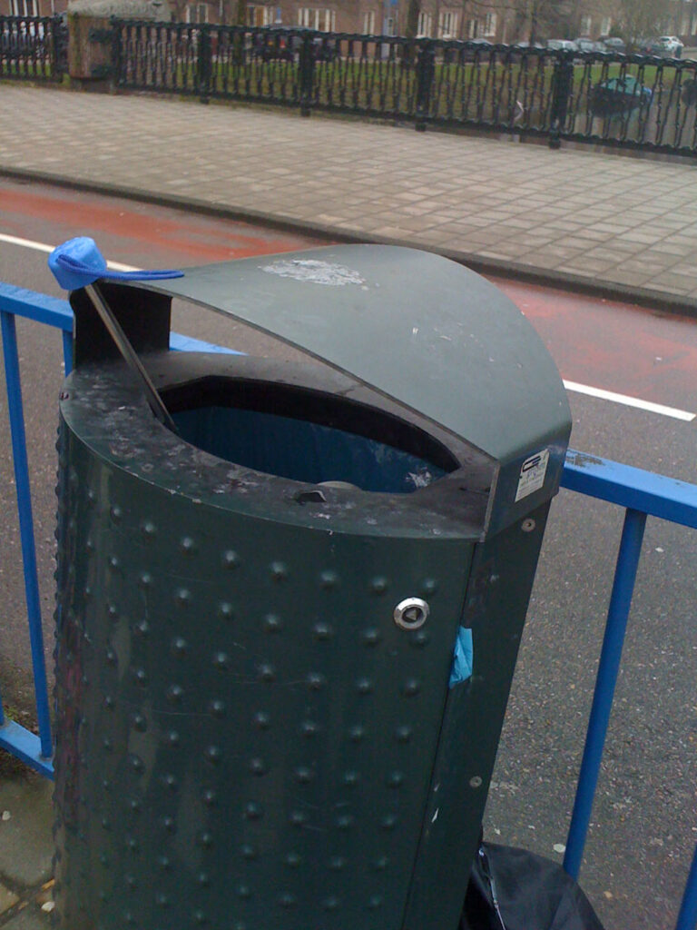 One brolly with blue handle in a bin and one black brolly next to it. Bin is on a tram stop which it self is on a Amsterdam bridge.