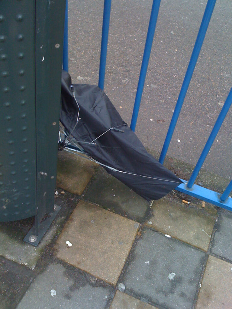 One brolly with blue handle in a bin and one black brolly next to it. Bin is on a tram stop which it self is on a Amsterdam bridge.