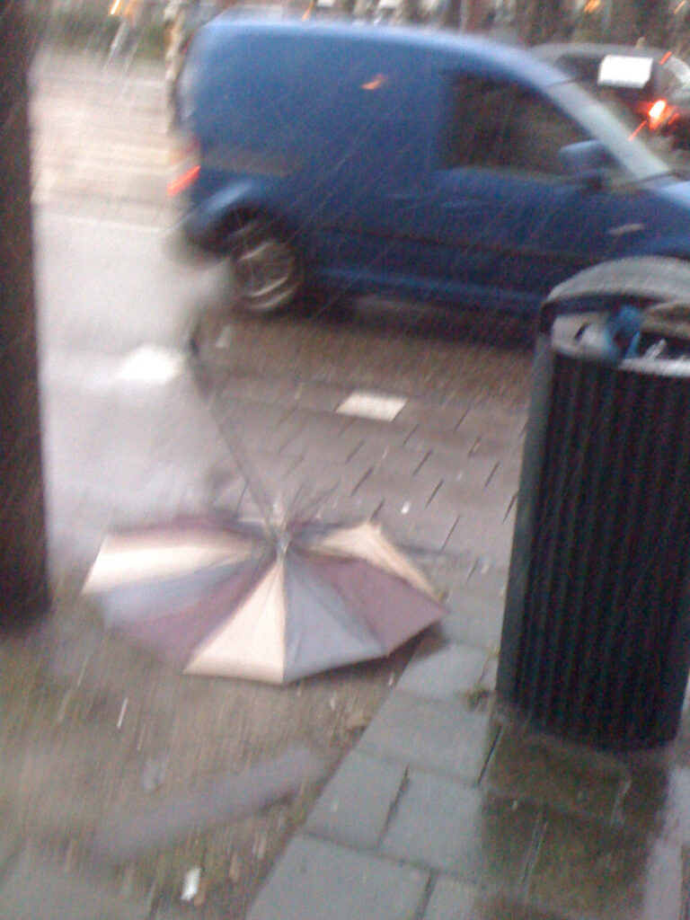 Brolly between tree and bin, all panels are flat on the ground. Tri colour fabric, upside down. Rain poors.