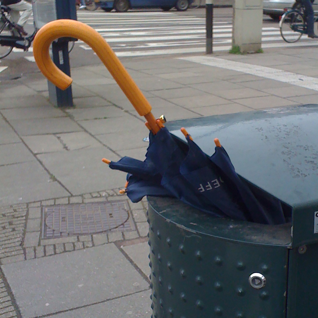 Classic brolly binned on a classic way. Handle and a bit of shaft and canopy sticks out of the bin. Bin is on a street corner next to the road and near a crosswalk.