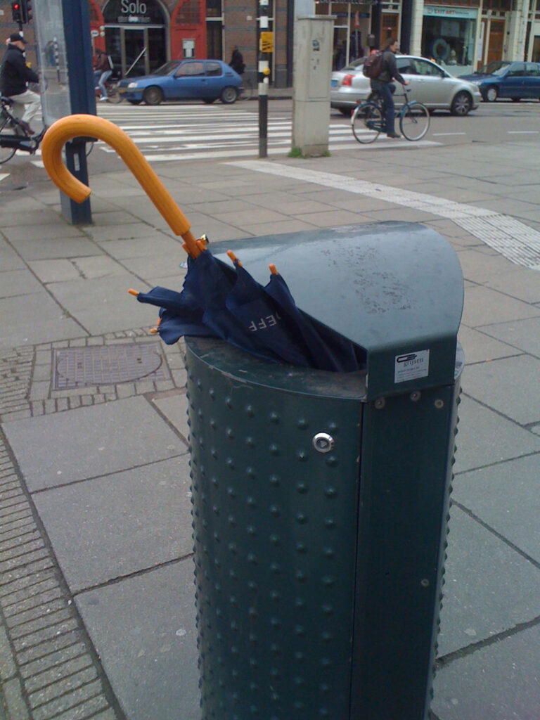 Classic brolly binned on a classic way. Handle and a bit of shaft and canopy sticks out of the bin. Bin is on a street corner next to the road and near a crosswalk.