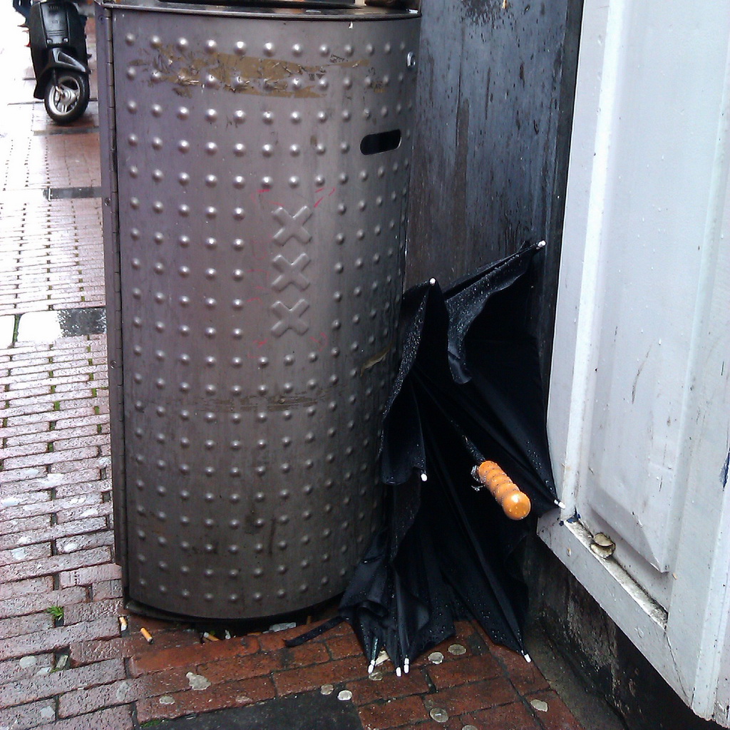 A brolly (black with wooden straigt handle) stuck betweel Bin and wall. Bin is in a shopping street. The street is wet.