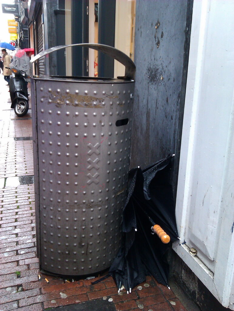 A brolly (black with wooden straight handle) stuck between Bin and wall. Bin is in a shopping street. The street is wet.