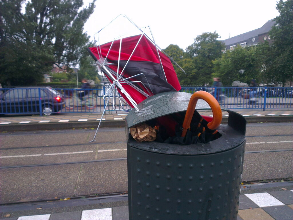 Two binned brollies. One classic binned, wooden handle and shaft and a bit of dark blue canopy sticking out. The second  one completely ruined top sticking out, black and red (Amsterdam) canopy mostly torn off the brolly ribs. Bin is on a tram stand on a bridge.