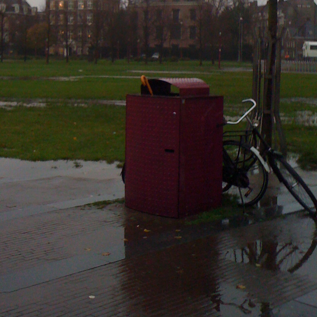 Big dark pink bin on the Museumplein. Square (bricks and grass, is very soaked of the recent rain. Brolly handle is just sticking out. Weather looks like you would imagine thinking about autumn.