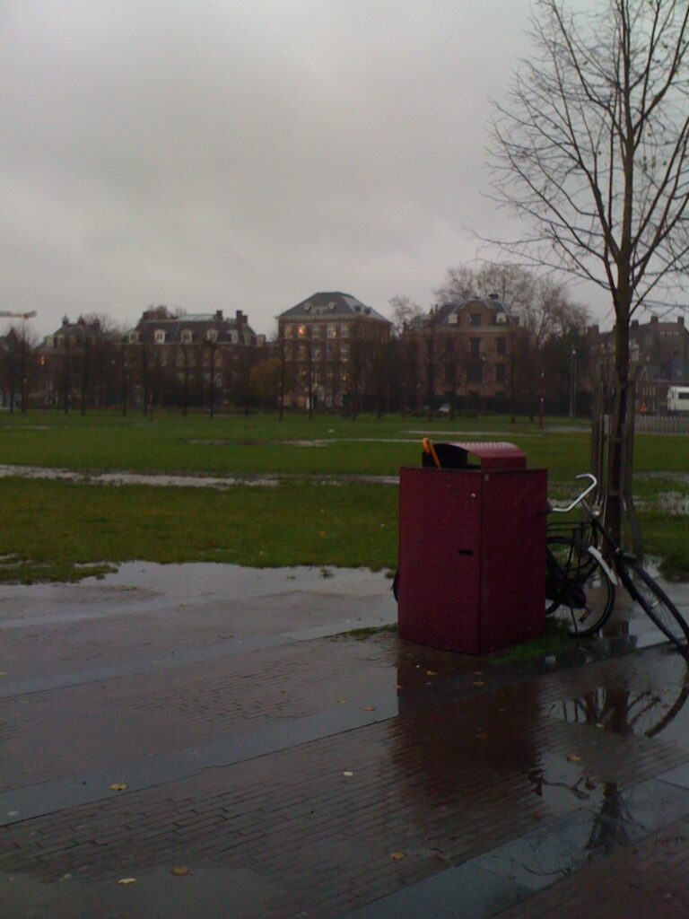 Big dark pink bin on the Museumplein. Square (bricks and grass, is very soaked of the recent rain. Brolly handle is just sticking out. Weather looks like you would imagine thinking about autumn.
