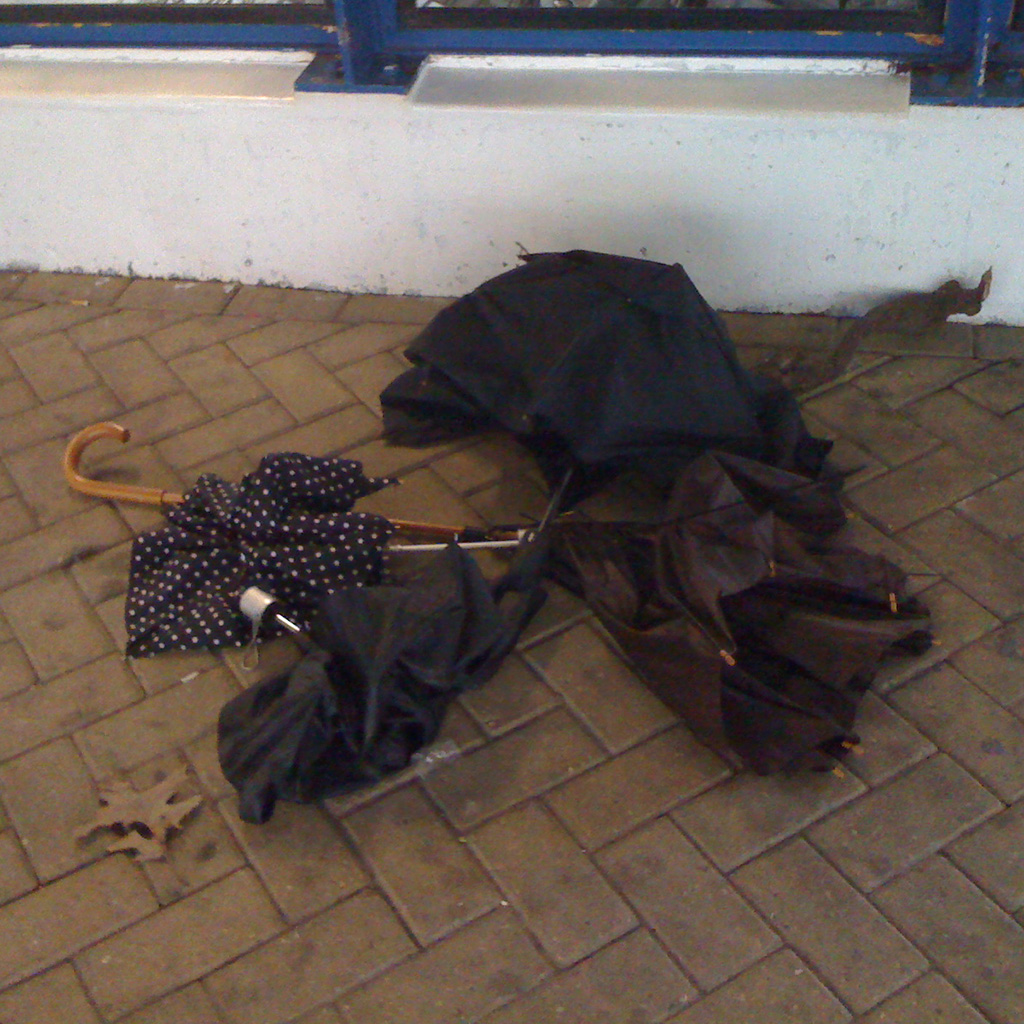 A gathering of four different kind of brollies lying on paving stones. Right next to a glass wall of a metro entrance. One brolly is brown with some red coloured pattern and a long shaft. This one looks like the most broken one. the two closest are small convertible types, one plain black and the other one black with white polkadots. The fourth one is also plain black and a bit bigger convertible one.