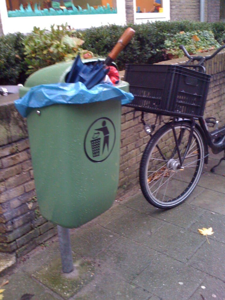 Classical binned brolly, blue canopy and wooden handle. Bin is a green smaller type plastic one. Mounted on a metal pole. Because the bin is quite full some other garbage is also visible. The bin is placed before a small brick wall next to a (school) entrance. The brick wall make a big planter in front of the school building. Next to the bin there is a bike with a crate.