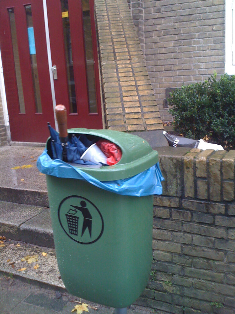 Classical binned brolly, blue canopy and wooden handle. Bin is a green smaller type plastic one. Mounted on a metal pole. Because the bin is quite full some other garbage is also visible. The bin is placed before a small brick wall next to a (school) entrance. The brick wall make a big planter in front of the school building. Next to the bin there is a bike with a crate.