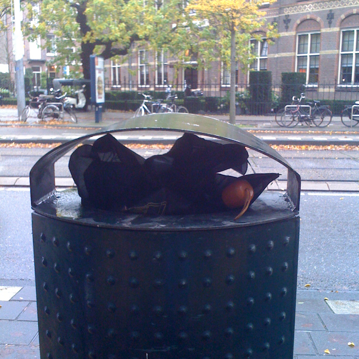 A folding brolly, black canopy and wooden (fake?) handle. Bin is a typical green Amsterdam bin. Background is some street and tram track. In one picture there’s van passing by, in another a tram.