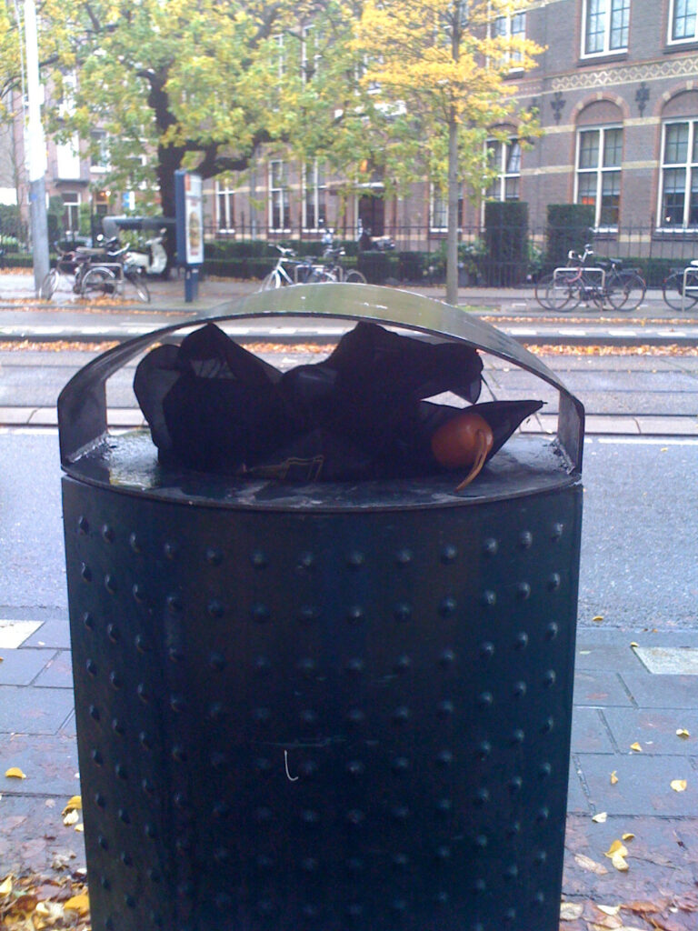 A folding brolly, black canopy and wooden (fake?) handle. Bin is a typical green Amsterdam bin. Background is some street and tram track. In one picture there’s van passing by, in another a tram.