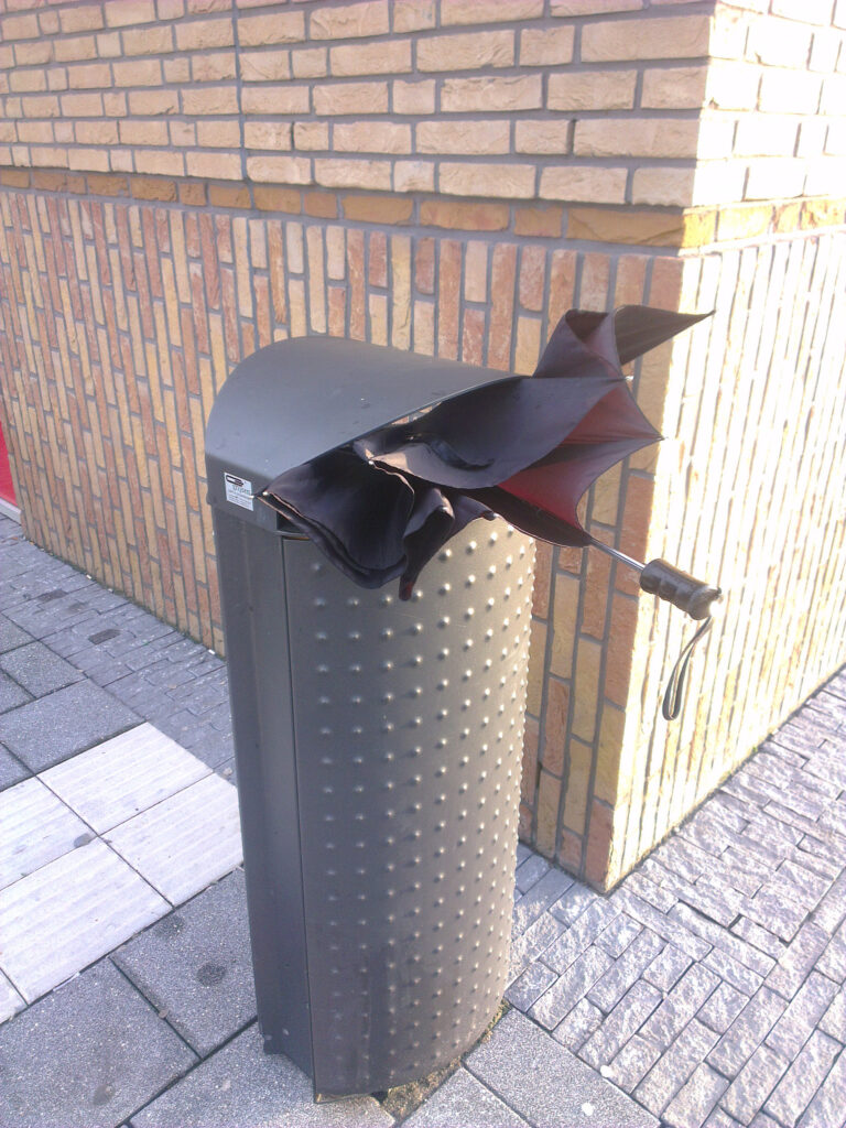 A folding brolly, black canopy and plastic handle. Smaal plastic loop attached to the handle. Bin looks quite new, grey matte painted. All set right next to a corner. Sunny on the right and shadow on the left of the corner. At the small view of the pavement you see three kinds of paving stones a.o. guide stones for blind and partially sighted people.