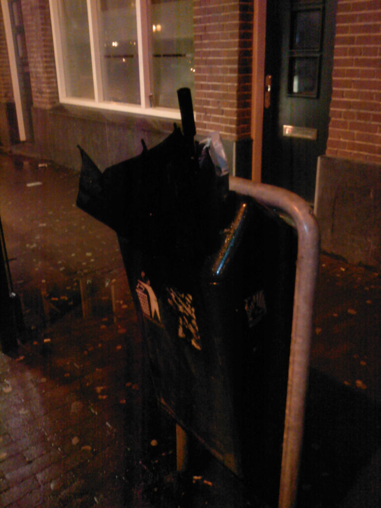 Real classic one, a black long brolly in a city bin. Street still wet from the rain. It’s nighttime but lots of city lightning so all is visible enough. Building in the background looks like it’s an office or something like that. 